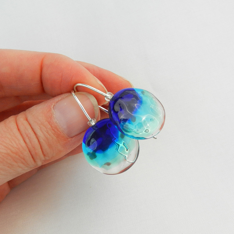 Silberne Ohrringe mit Glasperle in Bau und Türkis. Runde handgefertigte Glasperlen als Ohrhänger, wie Meeresschmuck. Unikate vo schmuckes Glas. in der Hand